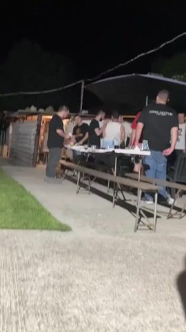 A nighttime outdoor shot shows several men gathered around two long tables on a concrete patio. To the left of the patio is a patch of green grass. In the background, there's a small structure with a light-colored corrugated roof, and beyond that, trees are visible against the dark sky. A black canopy or umbrella is positioned above the tables. The men are casually dressed, mostly in t-shirts and jeans. Some are holding drinks or small objects. They appear to be socializing. One man in a black t-shirt and jeans is facing the camera, standing to the far right of the tables. Another man in a black t-shirt and grey pants is on the far left, looking down at something in his hands.