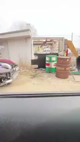 The video shows a red Chevrolet Corvette parked on a dirt lot. The front end of the car is incomplete, exposing the engine compartment. A man stands by the car, looking at his phone. The engine makes a loud noise, with a distinct turbo sound.  The surroundings appear to be a semi-industrial or commercial area, with small buildings and a lamp post visible.