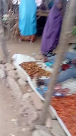 The video depicts an outdoor market scene. Initially, the ground with a white sack containing small brown objects is visible, along with blurred figures in the background, including individuals in purple and blue clothing. A person in a light patterned kaftan walks from right to left, followed by a small child in a light blue garment. The camera then pans to the right, revealing a bustling market area with multiple individuals, wooden tables, and makeshift stalls with thatched or tarp roofs. Two men are seated on wooden stools behind a table; one wears a dark outfit and a cap, and the other wears a light green outfit. They appear to be conversing, with the man in the dark outfit looking off-camera. Bags and other items are visible on the tables and around the stalls.