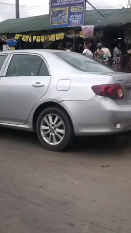 A silver car is parked on the side of a street, facing the viewer from the right of the frame. The car is partially obscuring a busy market scene behind it. The market appears to be an open-air street market with numerous stalls and vendors. People are seen walking, shopping, and interacting. Some individuals are carrying goods on their heads, a common practice in some regions. The stalls are covered with awnings and display various items for sale, though the specific items are not clearly identifiable. The ground in front of the market is paved, with debris and a drainage grate visible. The overall atmosphere suggests a lively and bustling commercial area. The sky is overcast, indicating cloudy weather.
