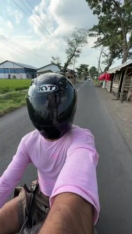 The video shows a first-person perspective of someone riding a motorcycle on a paved road. The rider is wearing a black full-face helmet with a "KYT" logo and a pink long-sleeved shirt. They are also wearing brown shorts. The motorcycle has a black bag attached to the back. The environment appears to be rural, with green fields and trees visible on the left side of the road. There are some buildings and structures in the background, including what looks like a greenhouse or covered farm area. The road is a two-lane asphalt road with white markings. The sky is cloudy. Another motorcycle is visible in the distance ahead.