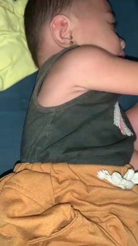 A close-up, side-view shot of a baby lying on its side. The baby is wearing a dark gray tank top and orange shorts. The baby's face is turned to the right, with its eyes closed and mouth slightly open. The baby's hair is dark and short. The background is blurred, but appears to be a bed or sofa with a blue surface and a yellow object partially visible. The lighting is dim.