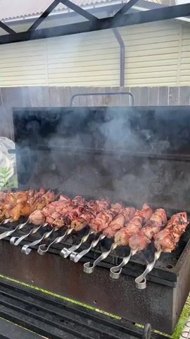 The video shows several skewers of meat cooking on a grill. The meat is raw and pink, and it is arranged in a line on the grill. There is smoke rising from the grill, and the meat is sizzling. A person's hand is visible in the upper left corner of the frame, holding a yellow spatula. The person is using the spatula to move the meat on the grill. The grill is outdoors, and there is a fence in the background. The fence is made of wood and is painted white. There is also a roof over the grill, which is made of metal. The video is shot from a low angle, looking up at the grill.