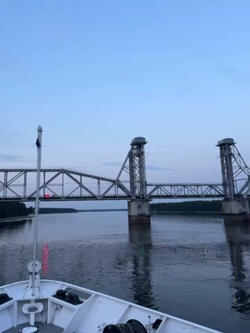 The video shows a wide river with a large metal bridge spanning across it. The bridge has two tall towers, and a section of the bridge appears to be raised, possibly for a boat to pass underneath. The water is calm, reflecting the sky and the bridge. The sky is a pale blue, suggesting it is either early morning or late evening. The foreground of the video shows the deck of a boat, with some equipment visible, including ropes and winches. The overall impression is of a peaceful scene during a boat journey.