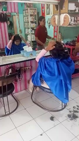 A woman sits in a salon chair, draped in a blue cape, facing a mirror. Her hair is dark and wavy. A man in a red shirt stands behind her, appearing to cut her hair. Another person, mostly out of frame, is also present. The salon has tiled floors, shelves of hair products, and promotional posters. Hair clippings are visible on the floor.