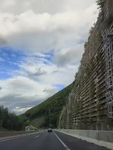 The video captures a view from a vehicle traveling on a highway. The highway has multiple lanes and is bordered by concrete barriers. To the right of the highway, a steep, tree-covered mountainside rises dramatically. The mountainside is reinforced with a series of concrete structures, creating a terraced effect, with visible metal supports and vegetation growing between the concrete sections. The sky above is partly cloudy, with patches of blue sky visible between the white clouds. The road ahead appears to be curving slightly. A dark-colored car is visible further down the highway, traveling in the same direction. The overall impression is one of a mountainous landscape with extensive infrastructure to stabilize the slopes alongside the road.