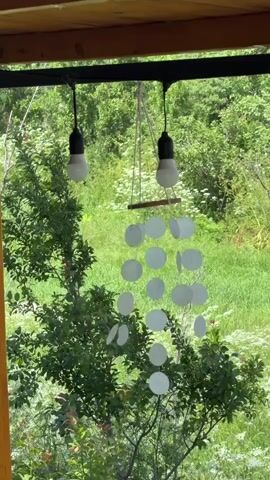 This short video captures a serene outdoor scene, likely on a porch or covered patio, overlooking a lush, green landscape. The primary focus is a handmade wind chime, composed of several circular, translucent discs suspended from a wooden crosspiece. The wind chime is hung from a dark horizontal beam, directly below two light bulbs that are also hanging from the beam. The light bulbs appear to be LED bulbs with a matte finish.

Behind the wind chime and light bulbs, a dense backdrop of green foliage dominates the scene. The trees and bushes are vibrant and full, suggesting a warm, perhaps summer, season. Small white flowers are visible scattered within the greenery, adding a touch of delicate detail. The overall impression is one of natural beauty and tranquility.

The weather appears to be pleasant, with ample natural light illuminating the scene. The gentle movement of the wind chime suggests a soft breeze. The setting in Vardablur, Armenia, implies a rural or semi-rural environment, characterized by abundant vegetation and a peaceful atmosphere.

No people are visible in the video. The activity taking place is passive, simply observing the gentle movement of the wind chime in the breeze and the beauty of the natural surroundings. The mood conveyed is one of peace, calm, and a connection with nature. There is no visible text within the video frame.