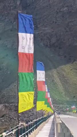 The video shows a line of colorful prayer flags on poles running along a bridge. The flags are blue, white, red, green, and yellow. The bridge appears to be in a mountainous region, with a rocky hillside in the background. The sky is bright, suggesting a sunny day. The flags are fluttering in the wind. The camera is moving forward, following the line of flags.