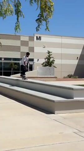 A young person in a white t-shirt and dark jeans performs a skateboarding trick on a set of concrete stairs. They ride down the stairs on their skateboard, jump off, and land on the ground. The skateboard then rolls away. The person then gets up and walks off-camera, turning to run in the opposite direction. The scene takes place outdoors on a sunny day in what appears to be a school or municipal parking lot. The background features a modern building with horizontal lines, trees, and a clear blue sky.