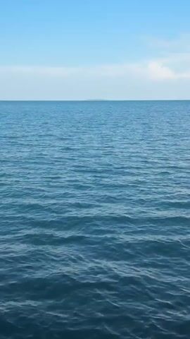 The video is a static shot of a vast expanse of blue water with gentle waves. In the distance, a very faint, flat landmass, possibly an island or a distant shore, is visible on the horizon. The sky above is a pale blue with a few wispy white clouds. The overall impression is one of calm and open space.