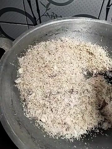 A cooking pan with a coarse, shredded white substance is being stirred by a dark utensil. The substance appears to be in the process of being toasted or dried. The pan is gray and sits on a stovetop. The background is a tiled wall with a subtle pattern. The lighting is dim, suggesting an indoor environment.