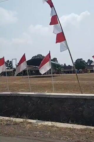 A row of Indonesian flags, red and white, are planted in the ground in front of a dry, grassy field. The flags are on poles, and several are waving in the wind. In the background, there are some buildings and trees, and a blue banner with text that appears to be in a foreign language. The sky is bright and cloudy. A dark concrete barrier is in the foreground, with some rocks and vegetation. A partial view of a scooter is visible at the bottom left of the frame.