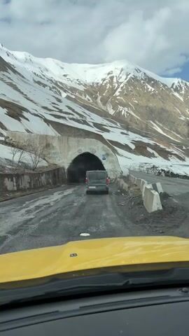 A dashcam view from a yellow car shows a gray van approaching a tunnel through a snow-covered mountain. The road is cracked and shows signs of disrepair. The sky is cloudy and bright. Height restriction signs are posted on the tunnel entrance, along with graffiti. The van is the only moving object visible.