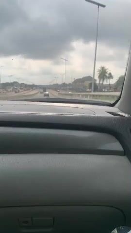 The video shows a first-person perspective from inside a moving car, looking forward through the windshield. The dashboard, which is light gray, occupies the bottom third of the frame. Through the windshield, a multi-lane highway is visible, with several cars driving in both directions. The sky is overcast with gray clouds. On either side of the highway, there are streetlights with what appear to be solar panels on top. Buildings, some appearing to be residential or commercial, line the sides of the road. A palm tree is visible on the right side of the frame in the distance. The car appears to be driving on the right side of the road.