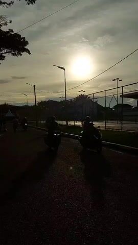 The video captures a street scene in Banda Aceh, Indonesia, during what appears to be the late afternoon or early evening, as indicated by the low angle of the sun and the warm, diffused light. The sky is partly cloudy, with the sun casting a bright glow through the haze. A tree with dark foliage dominates the left side of the frame, providing a strong silhouette against the sky.

The scene shows a road with several people walking and a few motorbikes. Some individuals are walking in groups, while others are more spread out. There's a general sense of movement and activity, though the pace seems relaxed. The subjects are silhouetted against the light, making it difficult to discern specific features, but they appear to be of various ages and are dressed casually.

In the background, there are structures that suggest a park or an outdoor recreational area, with what look like sports courts or fields enclosed by fencing. Lampposts are visible along the road, and some tents or canopies are set up in the distance, possibly for an event or market. The road itself is paved, and there are some wet patches, hinting at recent rain or maintenance.

The overall mood of the video is calm and serene, amplified by the soft lighting and the gentle movement of the people. The location cues, such as the style of the fencing and the general environment, along with the provided context, point to Banda Aceh, Indonesia. No visible text is present in the video.