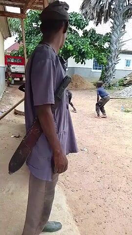 A man wearing a purple t-shirt, brown pants, and a dark cap stands with his back to the camera, facing away from a young boy. The man has a large, curved knife in a sheath tucked into his belt, and a small, dark object, possibly a gun, is holstered on his chest. The man's right hand is obscured by his t-shirt, but his left hand is visible. The boy, wearing a blue shirt and brown pants, stands with his back to the man, facing a pink object on the ground. The scene appears to be outdoors, with a dirt ground and a building in the background. There are also piles of dirt and debris, and a palm tree is visible to the right. A red truck is parked in the background to the left. The lighting suggests it is daytime.