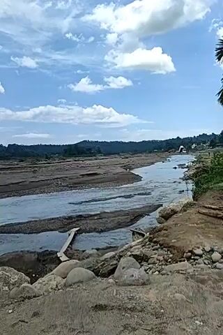 The video shows a wide, shallow river flowing through a rocky, somewhat arid landscape. The riverbed appears to be composed of sand, gravel, and larger stones. Several wooden planks are laid across parts of the river, possibly acting as a makeshift bridge or to divert water. In the foreground, there are large rocks and sandy soil. On the left side, there is a gabion wall constructed from wire cages filled with rocks, with two metal pipes extending upwards from it and into the water. In the background, the river continues to wind through the landscape, with trees and vegetation visible on the far bank. The sky is partly cloudy with a mix of white and darker gray clouds. The overall impression is of a natural, somewhat undeveloped environment, possibly undergoing some form of construction or water management project.
