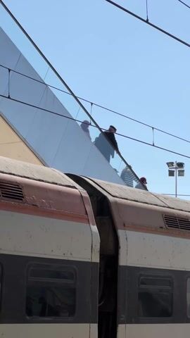 The video shows a double-decker train stopped at a station. The train is painted with white, grey, and orange stripes. A train worker is visible on top of one of the cars. People are walking on the station platform next to the train. The day is sunny with a clear blue sky. Overhead electrical wires run above the train and platform.
