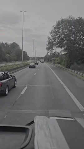 The video shows a point-of-view shot from inside a vehicle traveling on a highway. The road has multiple lanes, and several cars and a truck are visible ahead. The landscape on either side of the highway consists of trees and vegetation. The sky is overcast, suggesting a cloudy day. The dashboard of the vehicle is partially visible in the foreground.