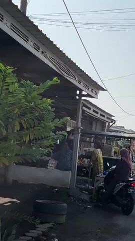 The video shows a street market scene. In the foreground, a person sits on a motorcycle. Behind them, a stall is set up with goods displayed. To the left, another stall has people interacting, and what appear to be food items are visible on tables. The background shows more market stalls and buildings, with a clear sky overhead. The overall environment appears to be a busy outdoor market.