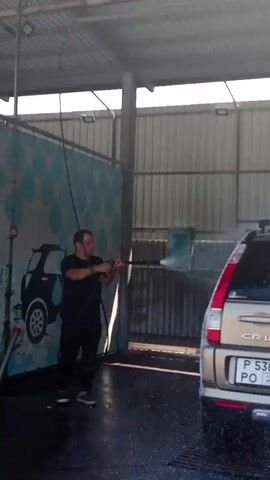 A man in a black t-shirt and black pants is pressure washing a gold Honda CR-V. He is holding a pressure washer wand with a black hose connected to it. The car is parked in an outdoor car wash bay with a corrugated metal roof and walls. There is a blue and white mural of a car on the back wall to the left of the frame. The ground is wet and soapy. The man is aiming the pressure washer at the rear passenger side of the car, and foam is building up on the car's surface. The car's license plate is visible and reads "P 538 PO". The car has a spare tire mounted on the rear.