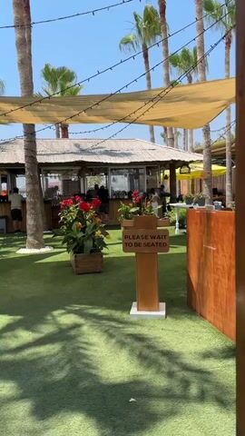 The video pans across an outdoor seating area with artificial turf. Palm trees are visible throughout the scene, with some strung with lights. A thatched-roof building, possibly a bar or restaurant, is in the background. A sign that reads "PLEASE WAIT TO BE SEATED" stands prominently on a wooden post in the foreground. In the background, people are seated at tables and walking around. The sky is clear and blue, suggesting a sunny day. Red and yellow flowers in a wooden planter add a splash of color to the scene.