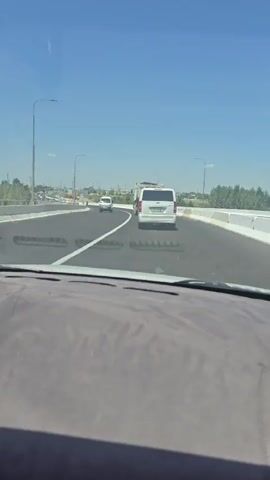 The video shows a view from inside a vehicle driving on a multi-lane highway. The road appears to be under construction or recently completed, with white lines marking the lanes and a concrete barrier along the right side. There are several vehicles on the road, including white vans and cars. In the distance, there are construction workers in orange vests and hard hats working near the side of the road, along with orange traffic cones. The sky is clear and blue, indicating a sunny day.