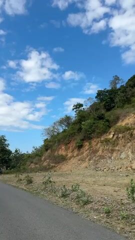 The video shows a steep, rocky hillside with sparse vegetation and a bright blue sky with scattered white clouds. A paved road is visible in the lower left corner of the frame. The hillside appears to be a quarry or an area where rock has been excavated, with visible layers of different colored stone. The camera pans slowly across the landscape, capturing the textures of the rock formations and the lush green foliage at the top of the hill. No people or animals are present in the video. The overall impression is one of a natural, rugged environment.