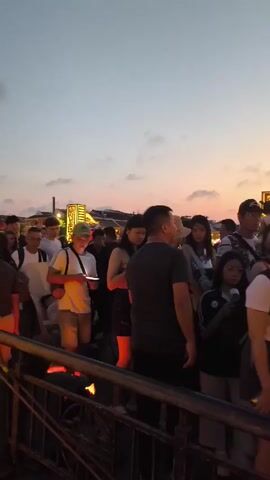 The video shows a crowded bridge at dusk, possibly in a tourist area. A diverse group of people is moving across the bridge. Many are young adults, dressed in casual clothes. Several individuals are looking at their phones. Along the bridge are tall, illuminated decorative structures resembling rectangular lanterns. The sky is a mix of orange and blue, indicating the time of day. The bridge has a metal railing and appears to be made of stone.