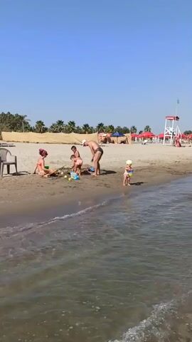The video shows a beach scene under a clear blue sky. In the foreground, gentle waves lap against the sandy shore. On the beach, a woman in a pink bikini sits on the sand, facing away from the camera, near some toys. Two children, a boy and a girl, are playing in the sand with the woman. A man, wearing dark swim trunks, stands near the children, interacting with them. A young child in a white hat stands by the water's edge. In the background, palm trees and beach umbrellas create a resort-like atmosphere. A lifeguard tower with a red and white structure stands prominently on the right side of the beach. The overall scene depicts a family enjoying a day at the beach.