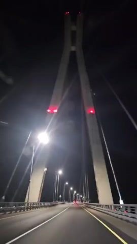 A first-person perspective video of driving on a cable-stayed bridge at night. The video starts with a view of the bridge's tall, concrete towers with red lights on top. The road ahead is well-lit by streetlights, and there are a few cars visible in the distance. As the car moves forward, hands briefly appear in the frame, obscuring the view of the bridge cables. The video continues to show the road and the illuminated bridge structure.