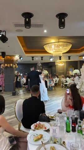 A wedding reception is underway. A couple, the bride in a white dress and the groom in a black suit, are dancing in the center of the room, illuminated by overhead lights. Guests are seated at tables, some observing the dancers, one woman in a red top is filming the couple with her phone. The room is decorated with floral arrangements, a large chandelier hangs from the ceiling, and spotlights are positioned throughout the space.
