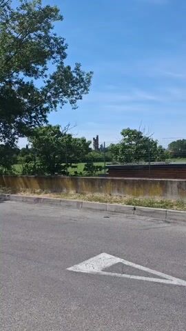 The video is a close-up shot of a tall, partially ruined tower, viewed through a wire fence. The tower is made of dark stone, with visible windows and architectural details. It is partially obscured by trees and bushes. The background is a bright blue sky with some clouds. In the foreground, there is a concrete wall with some weeds and a paved road with a white arrow painted on it.