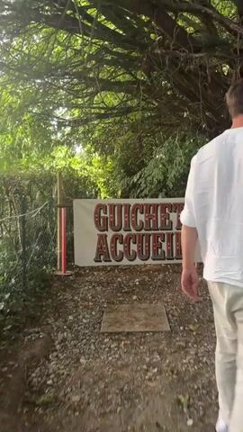 The video opens with a sign that reads "GUICHET ACCUEIL" in large red and yellow lettering, set against a white banner. The sign is positioned outdoors, likely at an entrance or information point, with a gravel path leading towards it. A person, seen from behind and mostly out of frame, is walking away from the camera towards the right.

The camera then pans slightly to the right, revealing a path that winds through a wooded area. The path is also covered in gravel. Further in the distance, a large, brightly colored circus tent is visible. The tent has alternating blue and yellow stripes and is decorated with stars. A banner with the words "CIRCUS & CO" is also prominent on the tent. People can be seen inside and around the tent, suggesting an event or performance is taking place. The overall environment appears to be outdoors, with trees and vegetation surrounding the area. The lighting suggests it is daytime.