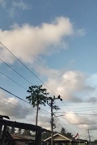 A short video shows a partially cloudy sky. Power lines and poles are visible in the foreground and middle ground, along with trees and some buildings. A red and white flag is also briefly visible. The video appears to be shot outdoors during the daytime.