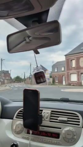 A first-person perspective video from inside a car shows a view of a road with buildings on either side. A roundabout is visible in the foreground, and a brick building with a grey roof is prominent on the right. The car is moving, as indicated by the blurring of the scenery. A black car dashboard with a phone mount and air vents is visible at the bottom of the frame. The sky is overcast with grey clouds. Outside the car, a beige baseball cap rests on the dashboard. The video appears to capture a drive through a town or village, with a mix of residential and possibly industrial buildings.
