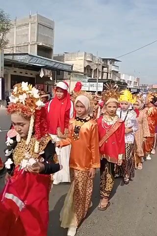 A group of children dressed in traditional clothing are walking down a street. The children are wearing colorful outfits, including head coverings and ornate headdresses. Some of the children are carrying flags. The street is lined with buildings and there are other people in the background. The weather appears to be sunny.