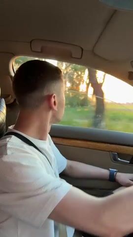 A young man with short hair is sitting in the driver's seat of a car and looking out the window. The sun is setting and casting a warm glow. He is wearing a white t-shirt and black shorts. He has a black bag slung across his body. The car's interior is visible, with a wooden trim on the door and a beige headliner. The man's hand is on the steering wheel.