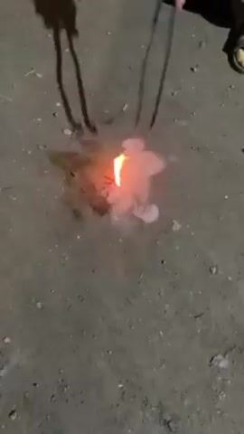 The video shows a small pyrotechnic device, possibly a firecracker or similar item, being ignited on a ground surface of dirt and small pebbles. The device emits a small flame and a trail of smoke before exploding in a bright flash and a cloud of white smoke. The remnants of the device are scattered across the ground. A person's legs and feet are visible in the periphery of the frame, implying someone is observing or interacting with the pyrotechnic. The environment appears to be an outdoor space, possibly at night.
