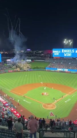 This is a shaky, high-angle shot of a baseball stadium at night. The scoreboard at the top right displays "ANGELS WIN! ANGELS WIN!". Smoke and fireworks are visible in the upper left corner and in the distance. On the baseball field, players in white uniforms are celebrating, some of them hugging. Spectators are seated in the foreground, looking towards the field.