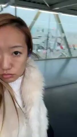 A woman with light brown hair and a white fur-trimmed coat is filming herself as she walks down a crowded airport corridor. The corridor has windows on the right side, and she is following a line of people. She appears to be looking directly at the camera.