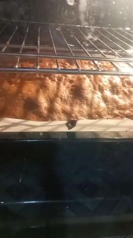 This is a close-up video, shot from the perspective of looking into a dark oven. The primary subject is a large, rectangular baked good, likely a cake or brownie, resting on a white baking sheet lined with parchment paper. The baked good is a rich, golden-brown color with a slightly cracked, bubbly surface, indicating it is fresh from the oven. The oven's wire rack is visible above the baked good. The background is dark, suggesting the oven interior.

The activity depicted is the baking process, or perhaps the final stages of baking. The visual cues of the hot oven and the browned surface of the baked good strongly suggest this. There are no people visible in the video.

Notable details include the texture of the baked surface, which appears somewhat uneven and porous. The lighting is bright and focused on the baked good, coming from within the oven. The timestamp at the beginning of the video indicates it was recorded at 00:00.

The location context provided is Oliveira do Bairro, Portugal. While there are no specific visual cues within the video to confirm this location, the timestamp and the general nature of home baking are universal. There is no visible text in the video.