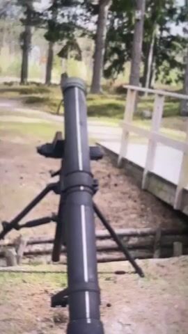 The video pans across a scene depicting a military mortar positioned on a tripod in a wooded area. The mortar is a large, dark cylindrical object with visible markings and attached to a tripod with metal legs. In the background, there is a dirt trench with wooden supports and a small wooden bridge. The environment is characterized by numerous tall trees with thin trunks, and the ground is covered in pine needles and dirt. The overall impression is a static display or demonstration of military equipment in a natural setting.