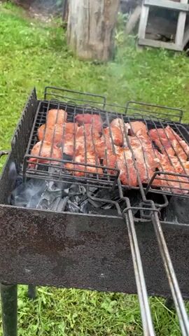 A video captures a stationary shot of a black metal barbecue grill with two grill grates on top, cooking what appears to be pork or some form of meat over hot coals. The background is a grassy outdoor area with some wooden objects and what looks like a shed. The smoke rises from the grill, and the meat cooks slowly. The camera remains still throughout the video.
