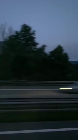 A side-view, panning shot taken from a moving vehicle, likely a car, captures a highway at dusk. The highway, with its distinct white lane markers, dominates the foreground. Beyond the highway, a dense line of dark green trees forms a backdrop. In the distance, a large, dark mountain range is visible, its silhouette stark against the fading light of the sky, which is a deep twilight blue. A single faint star can be observed in the upper portion of the sky. The trees show motion blur, indicating the speed of the vehicle. The overall atmosphere is serene and tranquil, conveying a sense of travel during the transition from day to night.