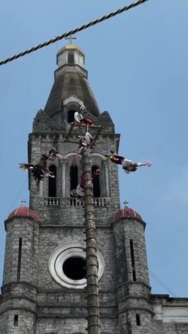 Against a blue sky stands a tall, gray stone church with a steeple. A tall wooden pole stands in front of the church. Attached to the top of the pole is a square metal frame from which four people in red and white costumes are suspended by ropes. The people are spinning in circles as the ropes unwind from the pole. A bird, perhaps an eagle, is also suspended from the pole. Two black electrical wires are visible across the top of the frame.