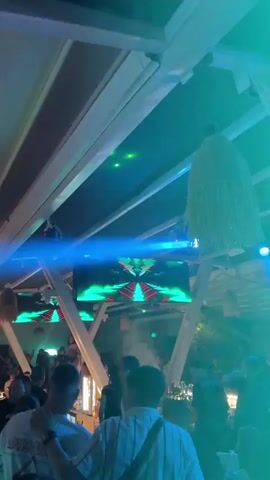 The video shows the interior of a crowded bar or club with a modern, open-air design. The ceiling is white with exposed beams and hanging decorative light fixtures resembling straw or grass. There is a large screen displaying abstract, colorful graphics behind the bar. Various lighting effects, including green laser beams and blue spotlights, illuminate the space. Many people are milling around, some near the bar area and others in the background. A haze or fog effect adds to the atmosphere.