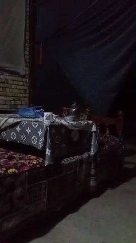 A low-angle, dark video captures a table with a patterned tablecloth, a teapot, and a mug. To the left, a brick wall is visible. A black tarp hangs in the background. The scene is dimly lit, with shadows obscuring details.