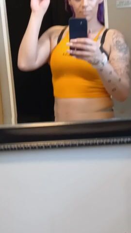 A person with purple hair is standing in front of a mirror taking a selfie. They are wearing an orange tank top and have visible tattoos on their left arm. They are holding up a strand of their hair with their left hand while holding a black smartphone in their right. The background includes a black curtain or door and a wooden shelf unit. The mirror has a black frame, and the wall surrounding it appears to be a light neutral color. The lighting seems to be indoor and slightly diffused.