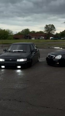 Two black Lada Priora cars are parked on an asphalt surface under a cloudy sky. The car on the left is a hatchback, while the car on the right is a sedan. The sedan is further away from the camera and has a gray wheel rim. There is a pool of water on the ground near the hatchback. The background features a couple of buildings, trees, and some other vehicles.